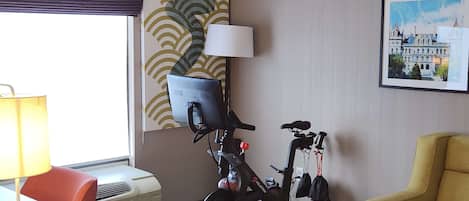Une chambre d’hôtel équipée d’un bureau, d’une chaise, d’un vélo d’exercice, d’un climatiseur, d’une lampe et d’une œuvre d’art au mur.