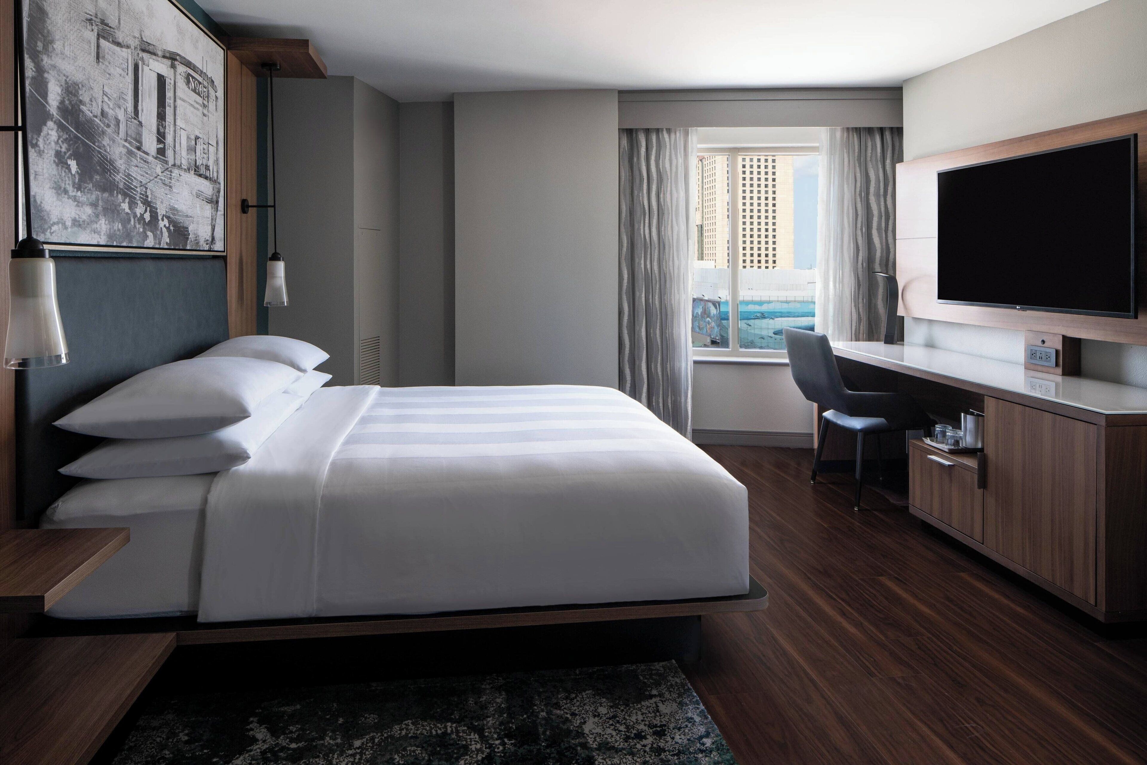 A modern hotel room with a large bed, a desk, a flat-screen TV, and a view of the city through the window.