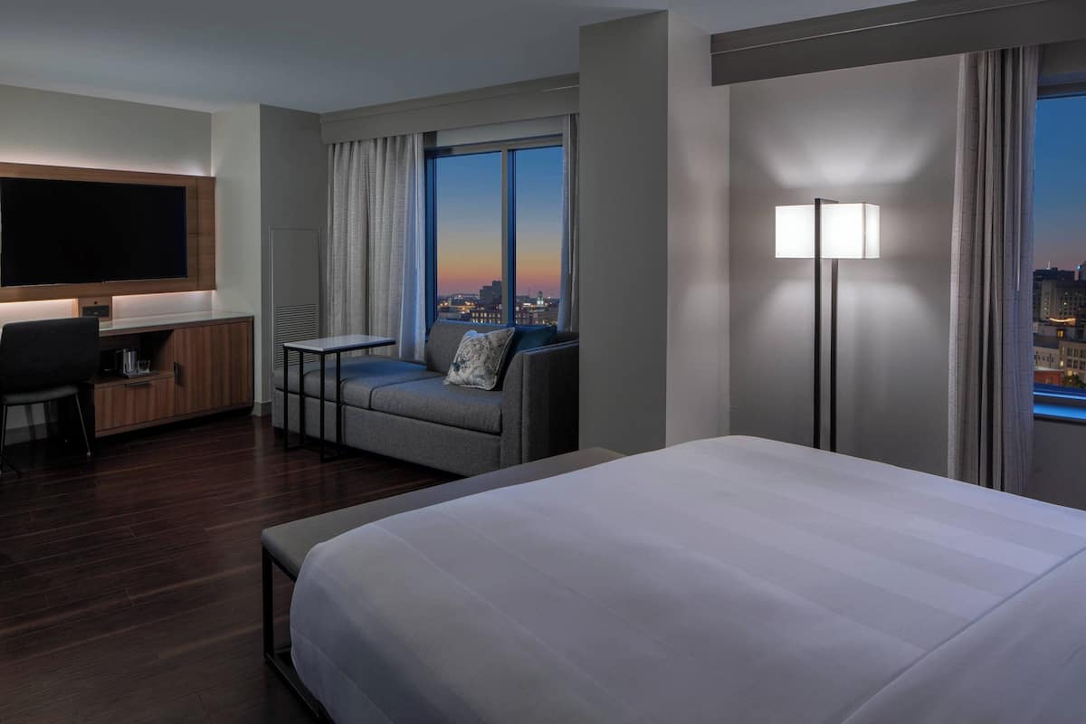 A hotel room with a large bed, a desk with a chair, a sofa with a view, and a TV mounted on the wall.