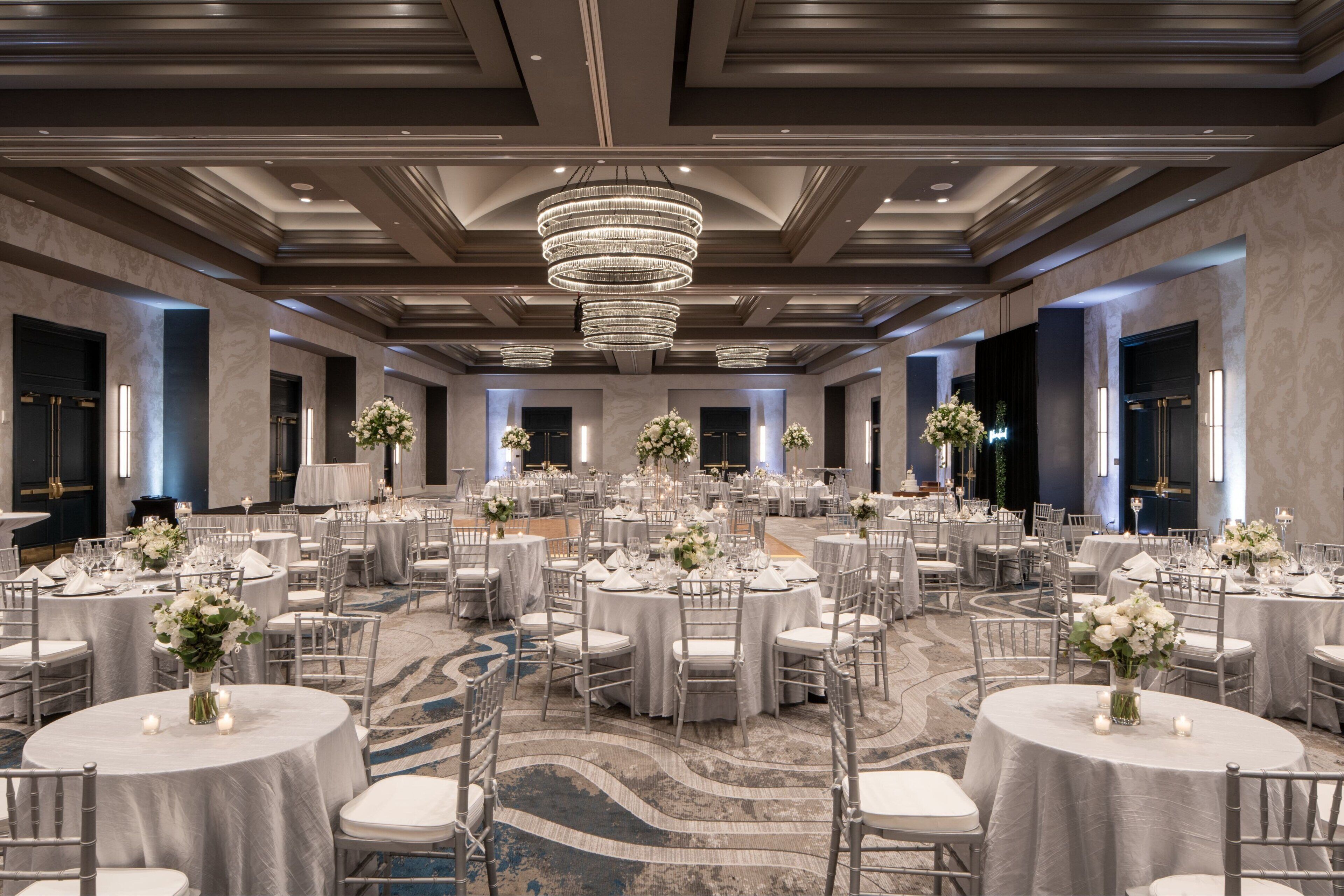 A large, well-lit banquet hall with round tables set for events, featuring chandeliers and floral centerpieces.