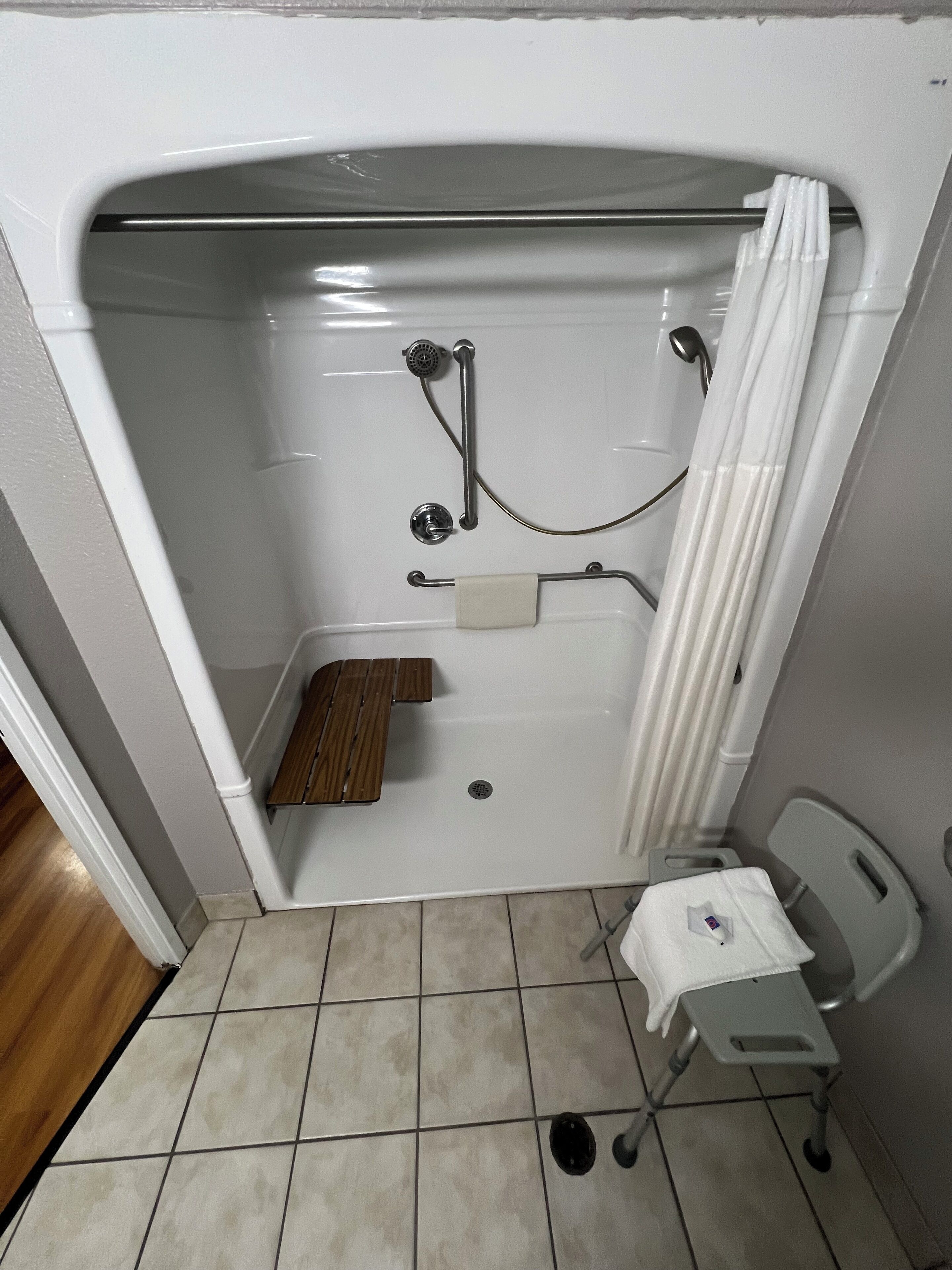 A small, enclosed shower area with a white shower curtain, a handheld showerhead, a handheld shower head, a small step stool, and a white towel on a small chair.