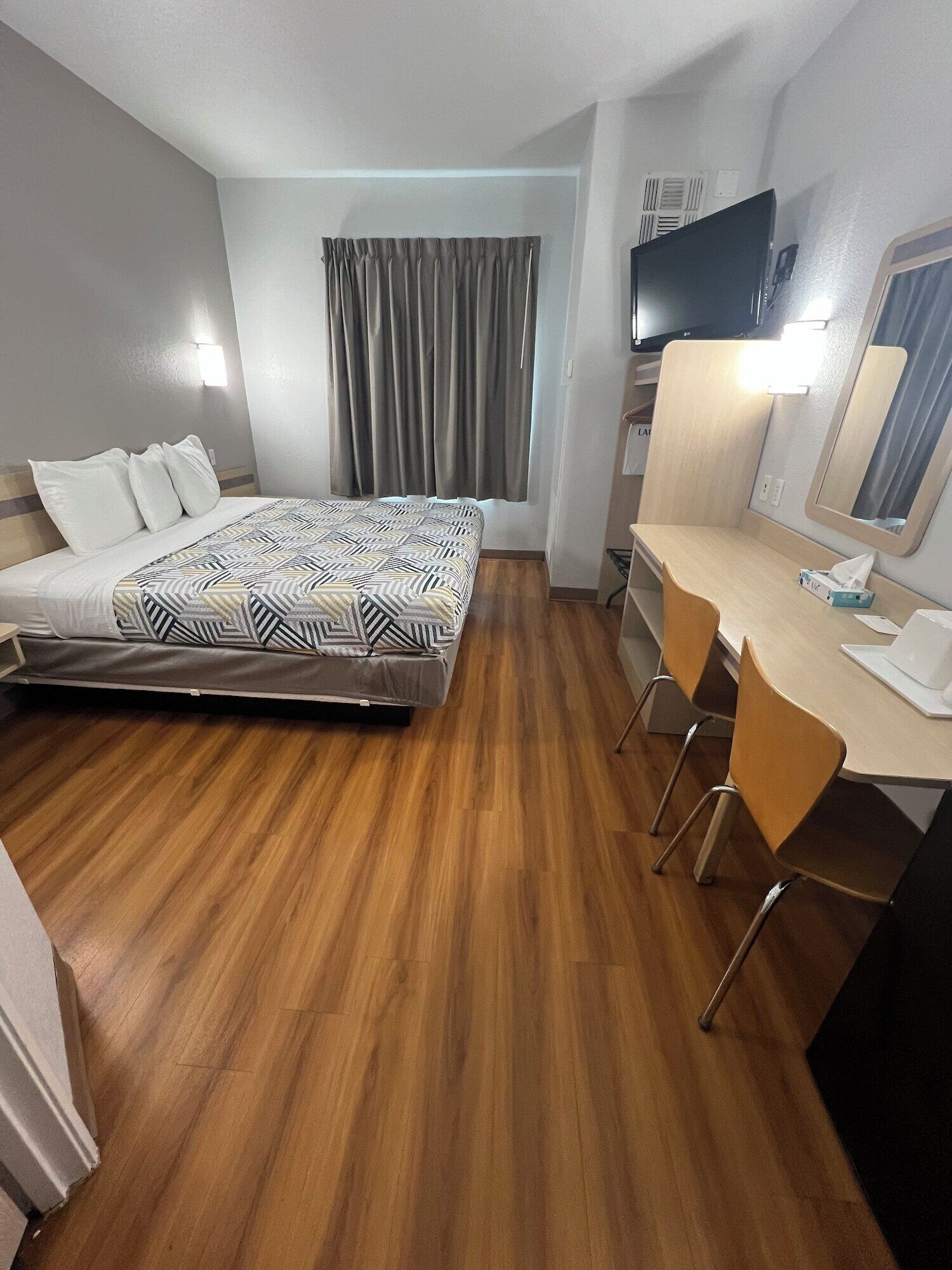 Una habitación de hotel con cama, escritorio con silla, televisor y ventana con cortinas.