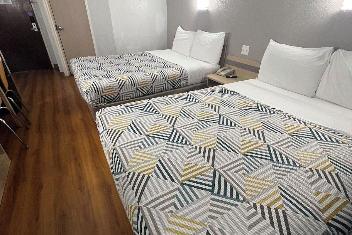 A hotel room with two beds, a wooden floor, a door, and a wall-mounted light fixture.