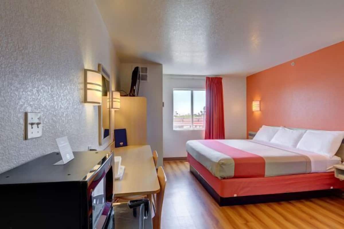 A hotel room with a bed, a desk with a chair, a window with curtains, and a wall-mounted lamp.