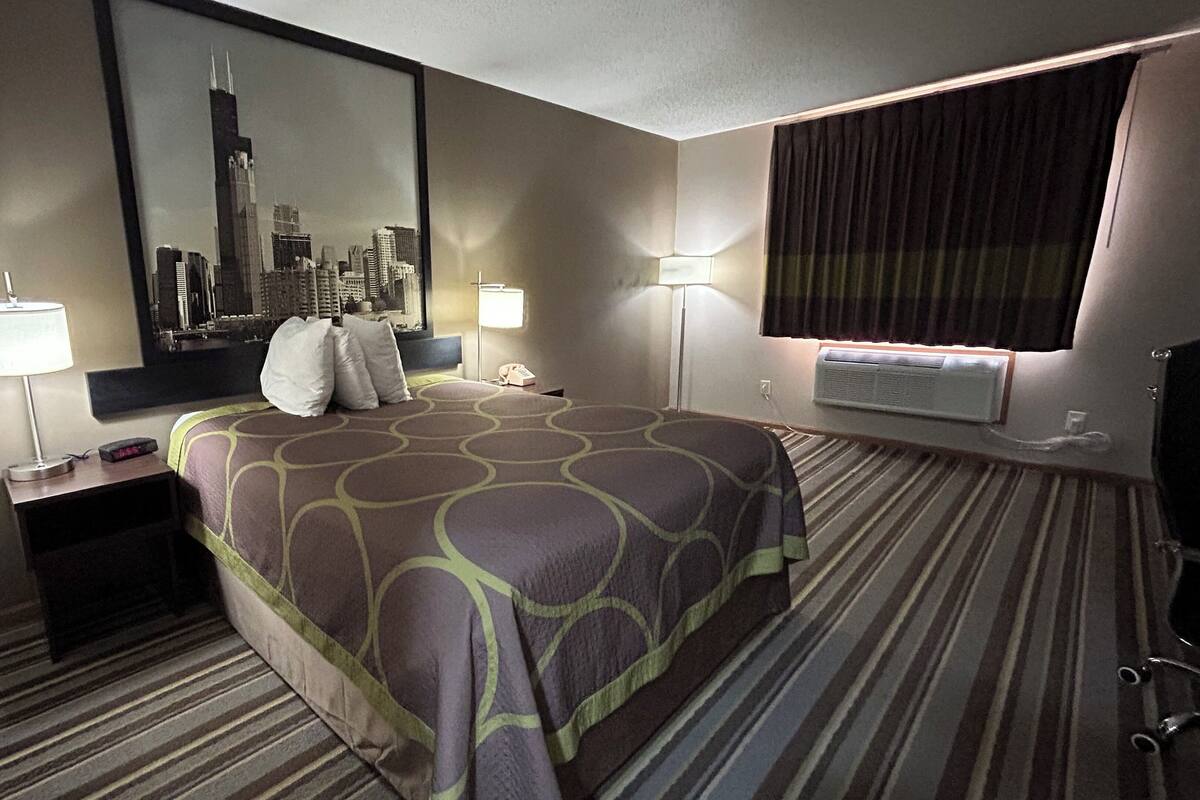 Una habitación de hotel con una cama grande, dos mesitas de noche con lámparas, un espejo grande, una ventana con cortinas y un equipo de aire acondicionado.