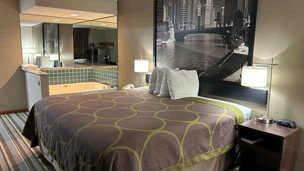 A hotel room with a large bed, a bathroom with a bathtub, a large mirror, and a cityscape view. - Super 8 by Wyndham Gilman (Gilman)