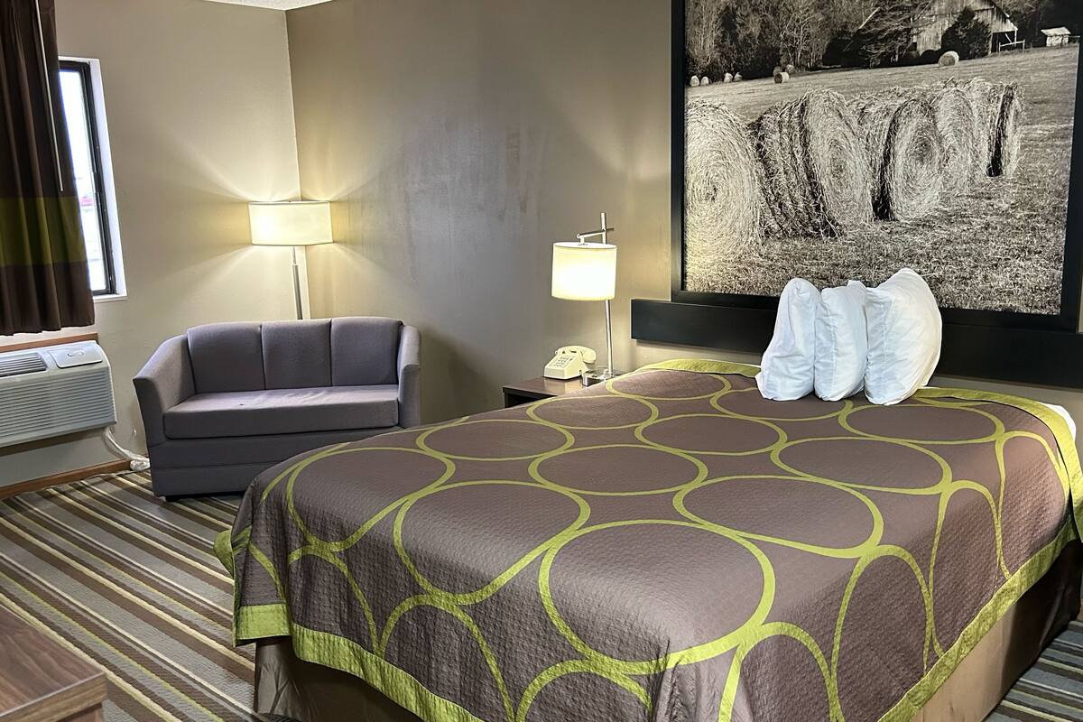 Habitación de hotel con cama, una silla, un escritorio, una lámpara y un cuadro en la pared.