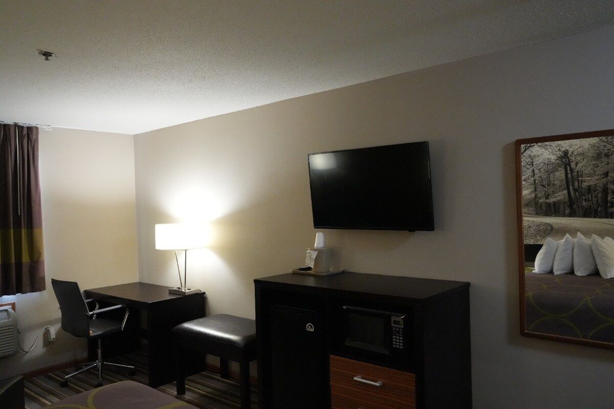 A hotel room with a desk, chair, television, microwave, and a painting on the wall.