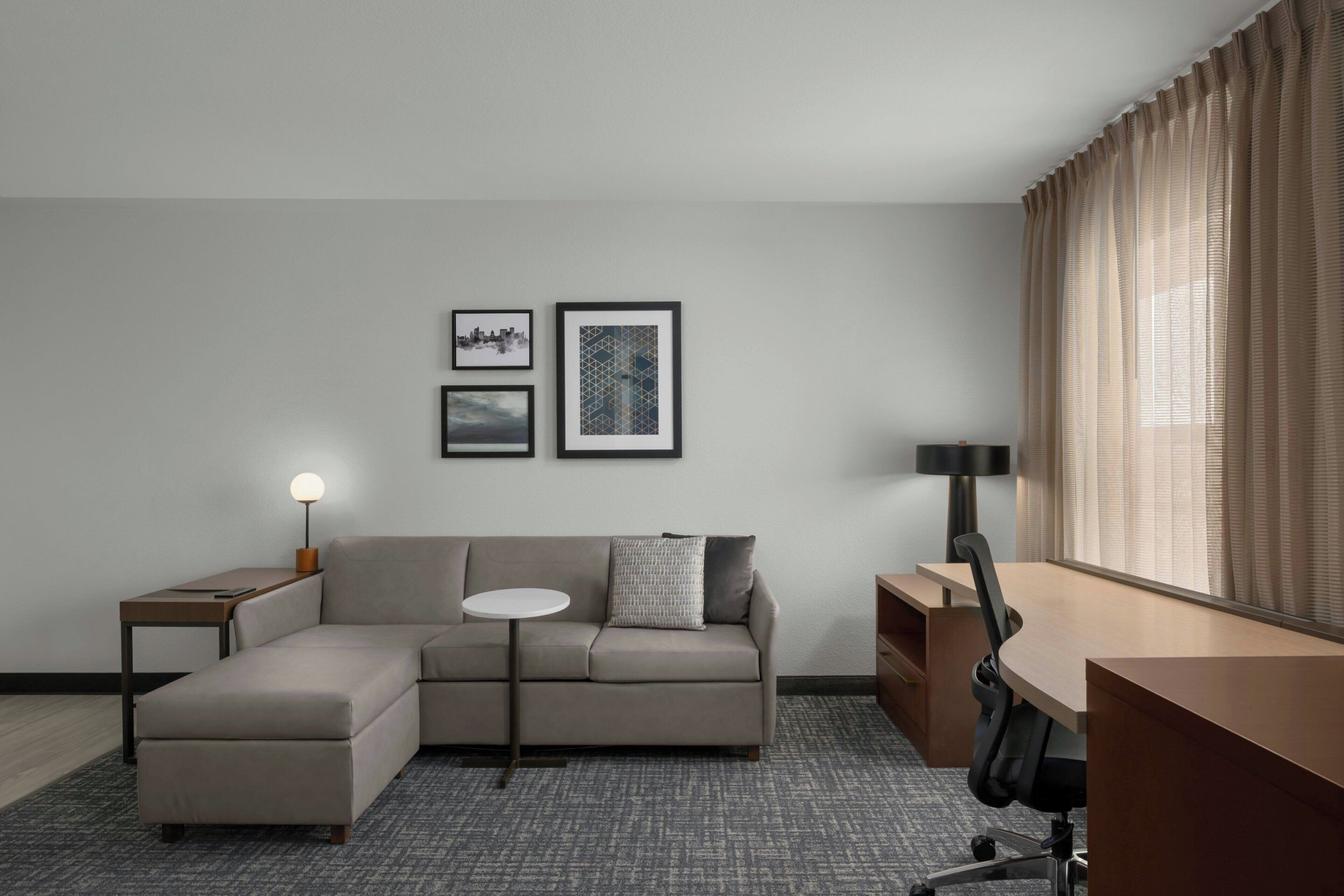 A modern hotel room with a sectional sofa, a small round table, a desk, and two framed pictures on the wall.