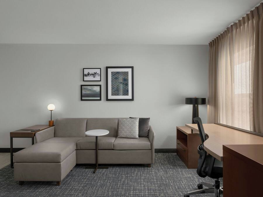 A modern hotel room with a sectional sofa, a small round table, a desk, and two framed pictures on the wall.