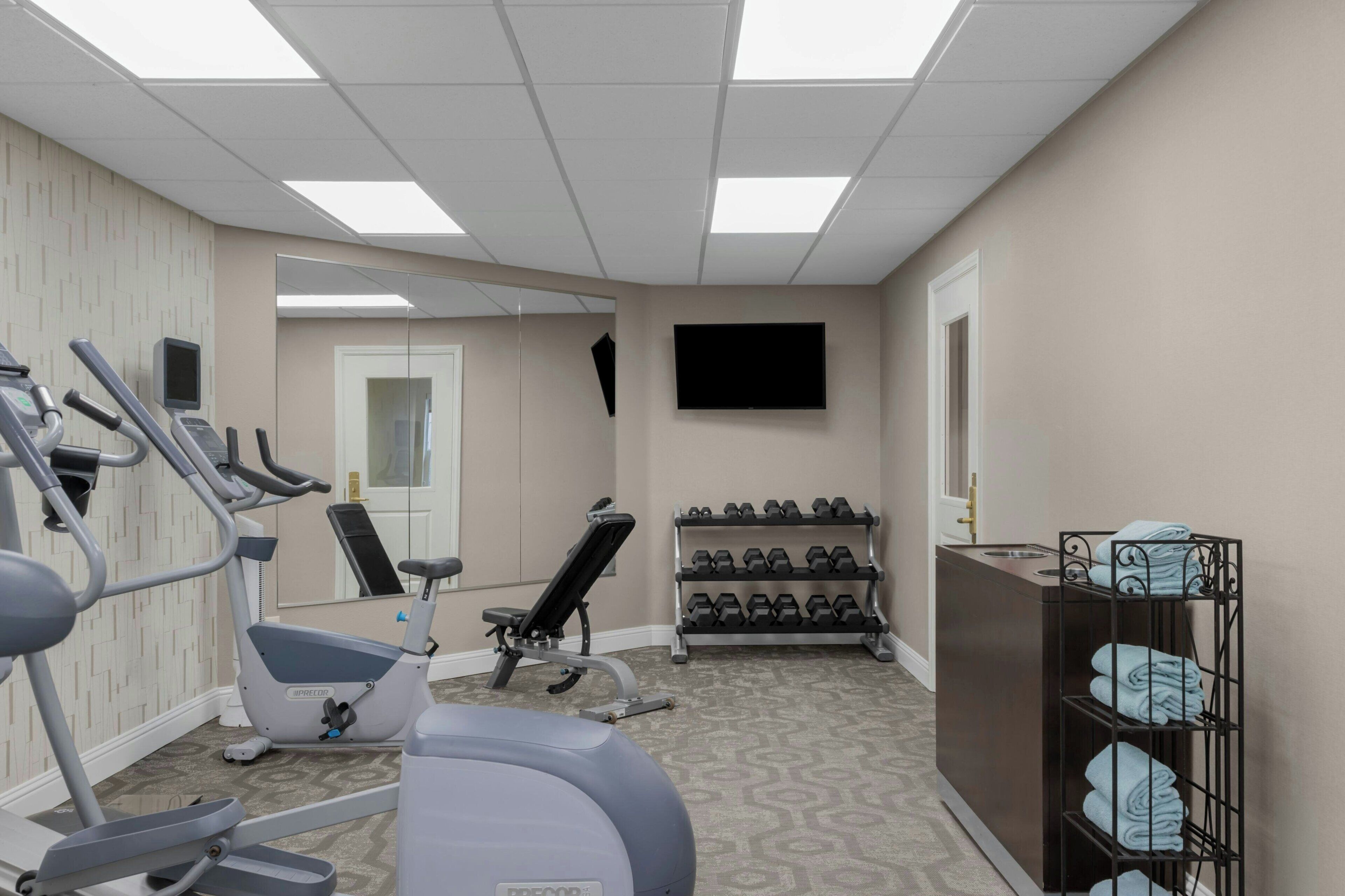 Un gimnasio con equipo de ejercicio, televisión, espejo y un estante con mancuernas.