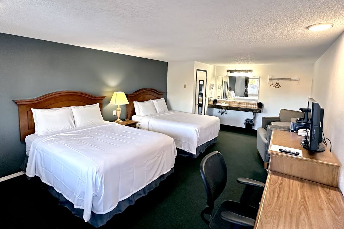 A hotel room with two beds, a desk with a computer, a chair, a mirror, and a bathroom with a sink and mirror.