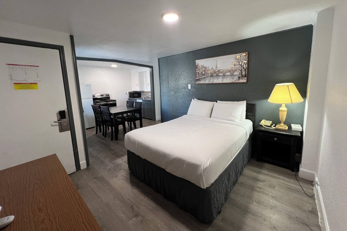 A hotel room with a bed, a desk, a chair, a microwave, a sink, and a painting of a bridge.