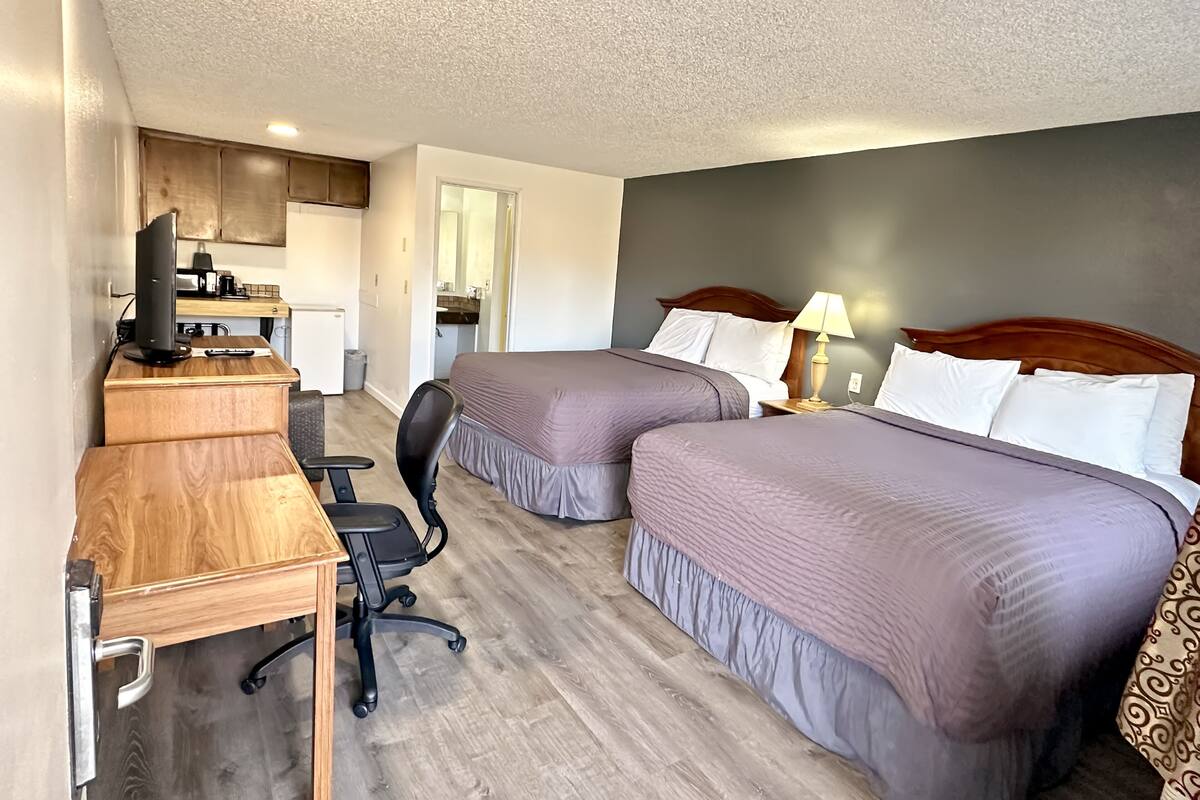 A hotel room with two beds, a desk with a computer, a chair, and a kitchenette in the background.