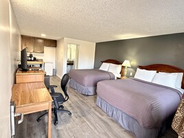 Habitación de hotel con dos camas, un escritorio con computadora, una silla y una kitchenette al fondo.