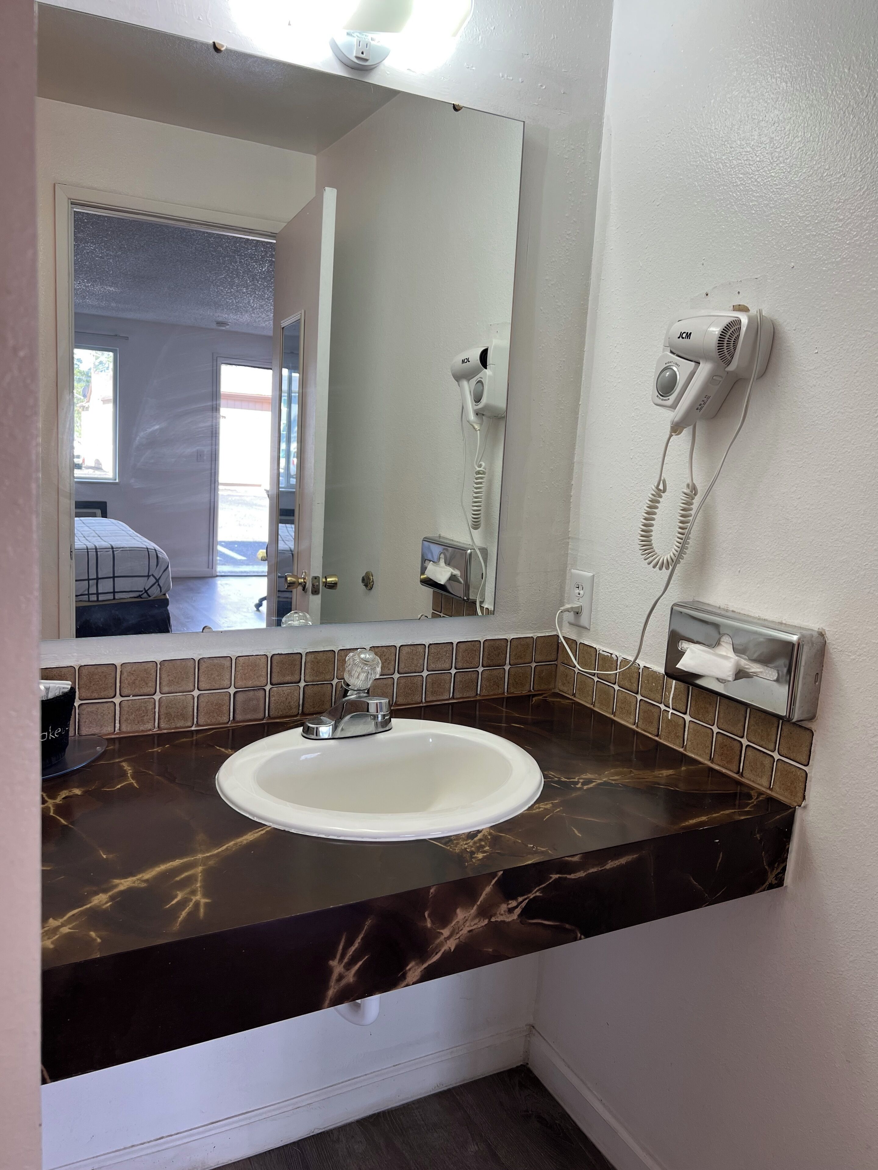 Uma casa de banho com uma pia branca, um espelho, um secador de cabelo e um dispensador de lenços.