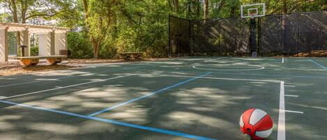 An outdoor basketball court with a basketball, surrounded by trees and a covered seating area.