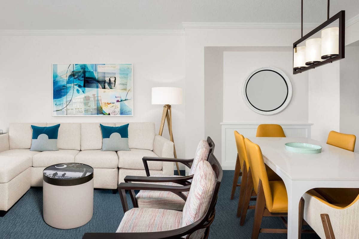 A modern living room with a sofa, armchairs, a coffee table, and a dining table with chairs. There is a painting on the wall and a floor lamp.