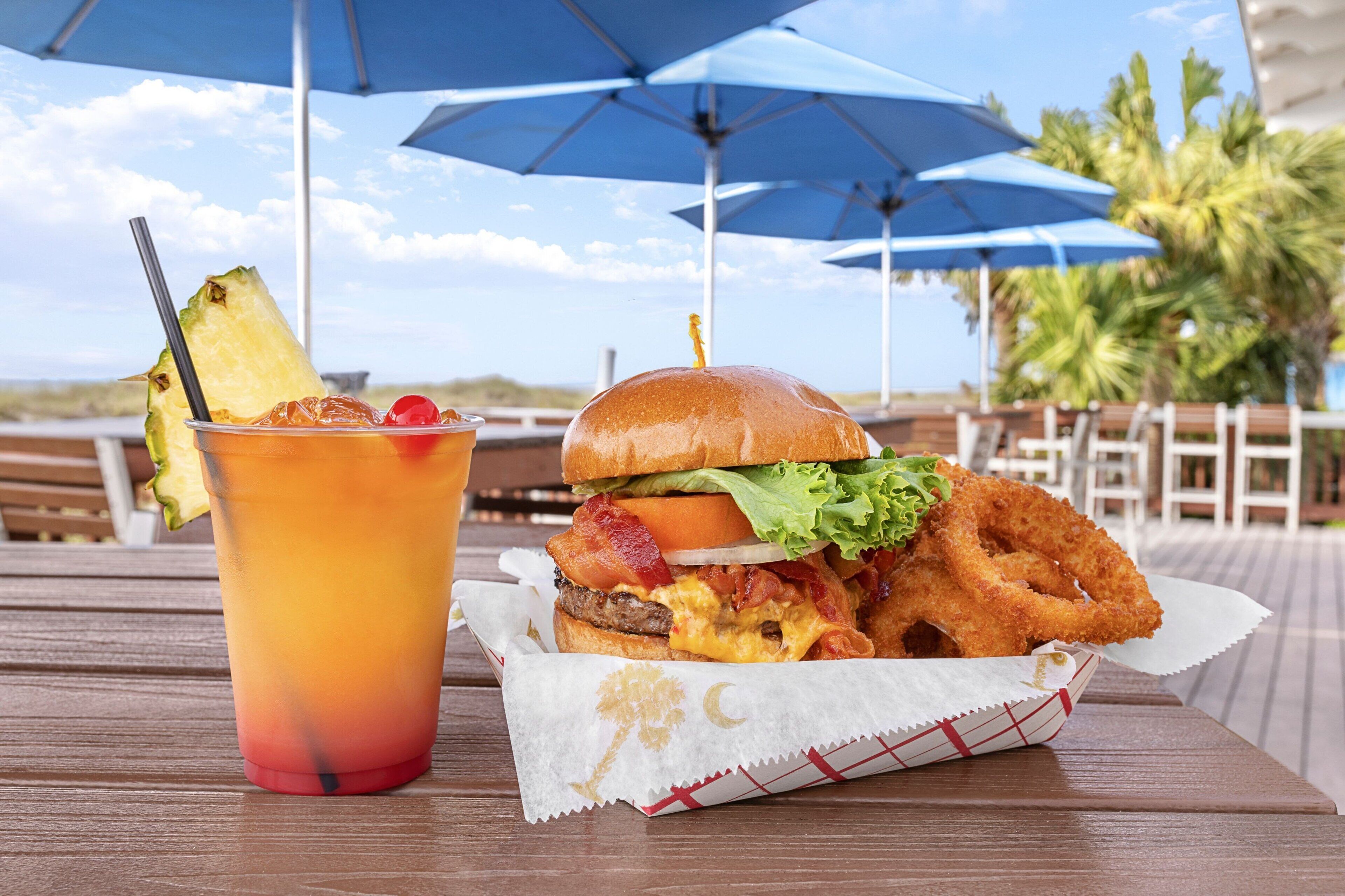 A meal consisting of a burger, fried items, and a drink with a pineapple slice on a wooden table under blue umbrellas.