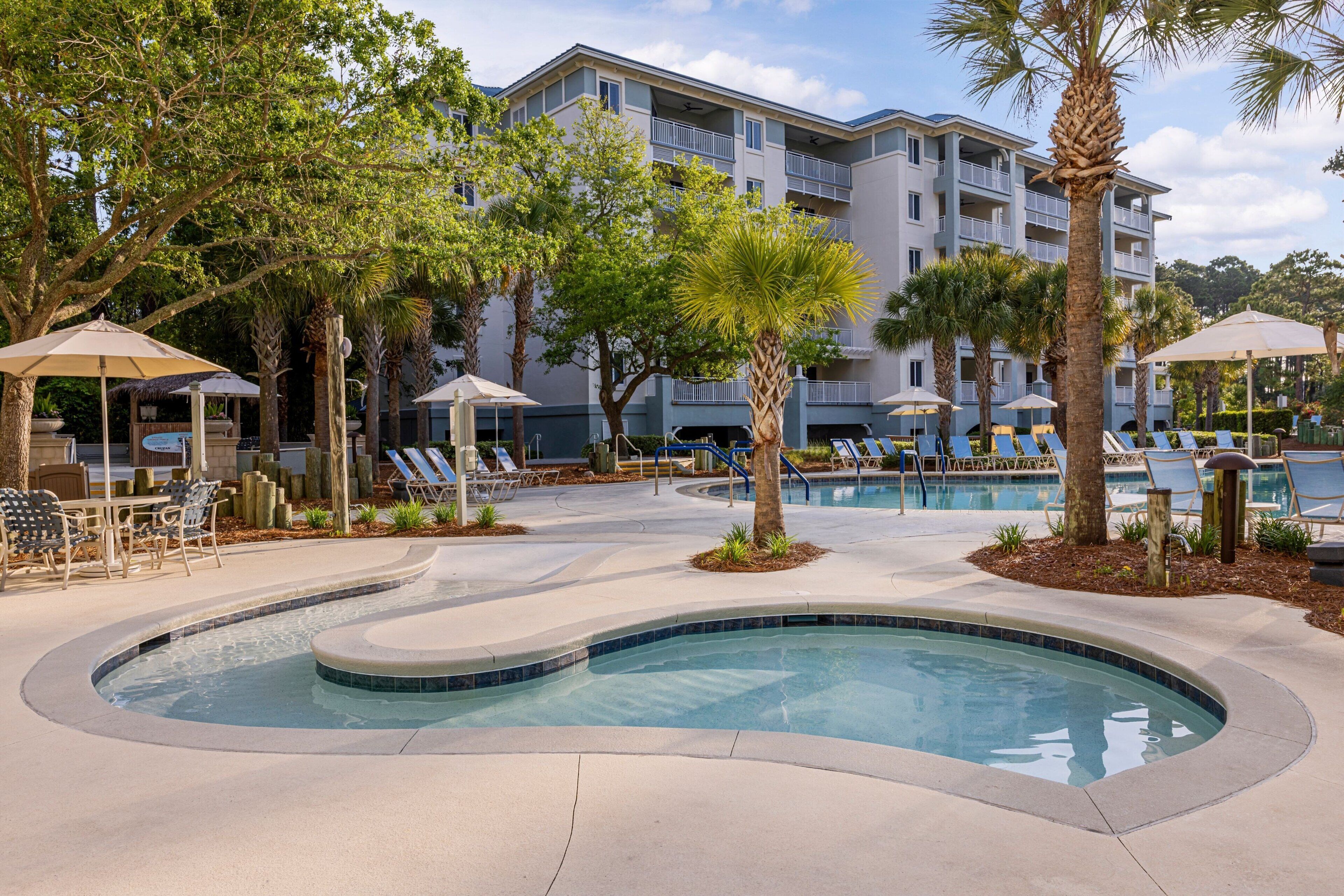 A pool area with lounge chairs and umbrellas, surrounded by palm trees and a building with balconies.