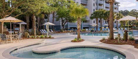 A pool area with lounge chairs and umbrellas, surrounded by palm trees and a building with balconies.