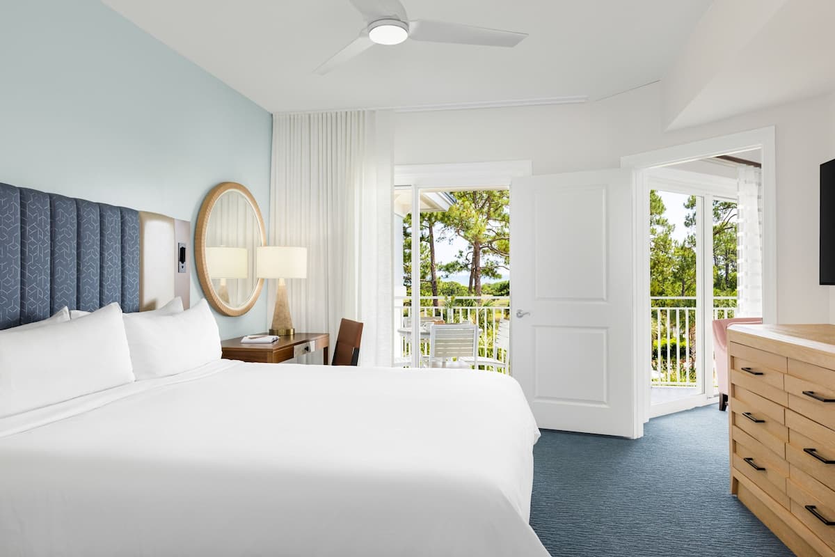 A hotel room with a large bed, a desk with a lamp, a mirror, and a dresser. There is a balcony with a view of trees and a white railing.