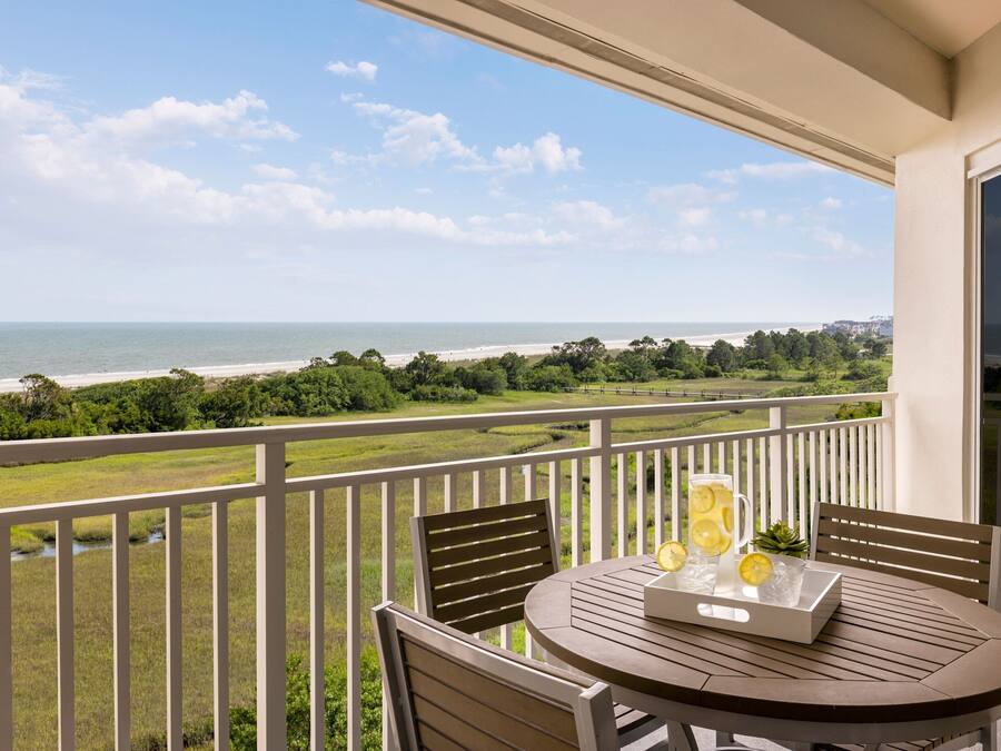 A balcony with a table set for two, overlooking a beach and greenery.
