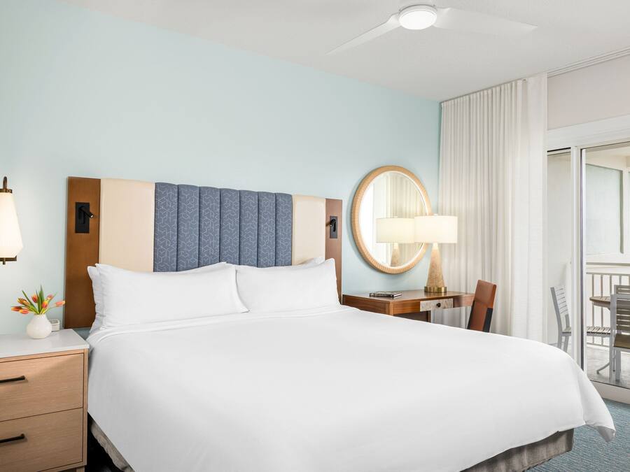 Habitación de hotel con una cama grande, una mesita de noche con lámpara, una pequeña mesa con una silla y vistas al exterior a través de una puerta corrediza.