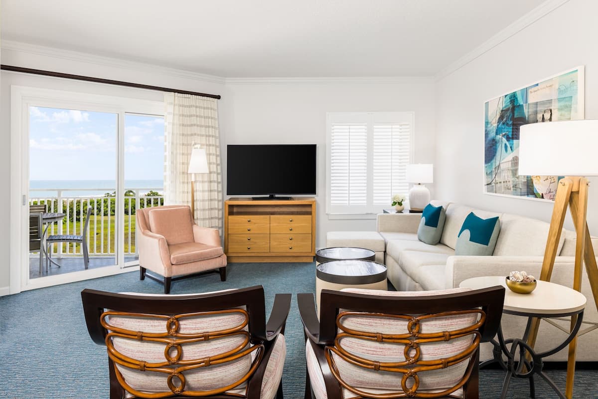 A living room with a television, a sofa, a chair, a small table, and a balcony with a view of the ocean.