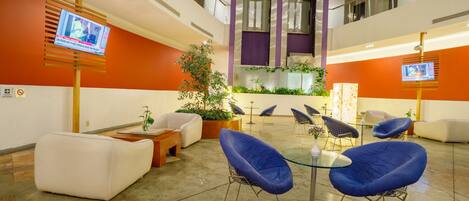 A modern lobby with a high ceiling, a central seating area with sofas and chairs, a coffee table, and a large plant.