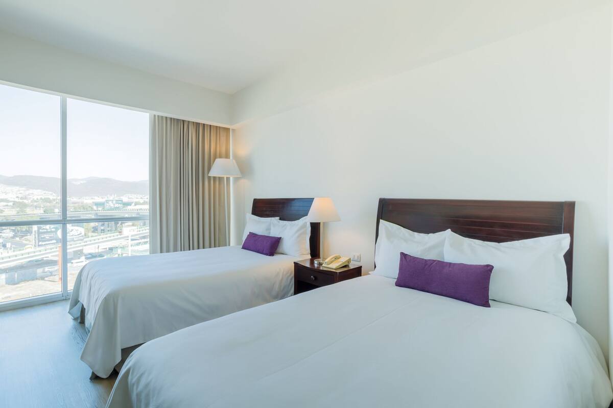 A hotel room with two beds, a large window with a view of the city, and a nightstand with a lamp.