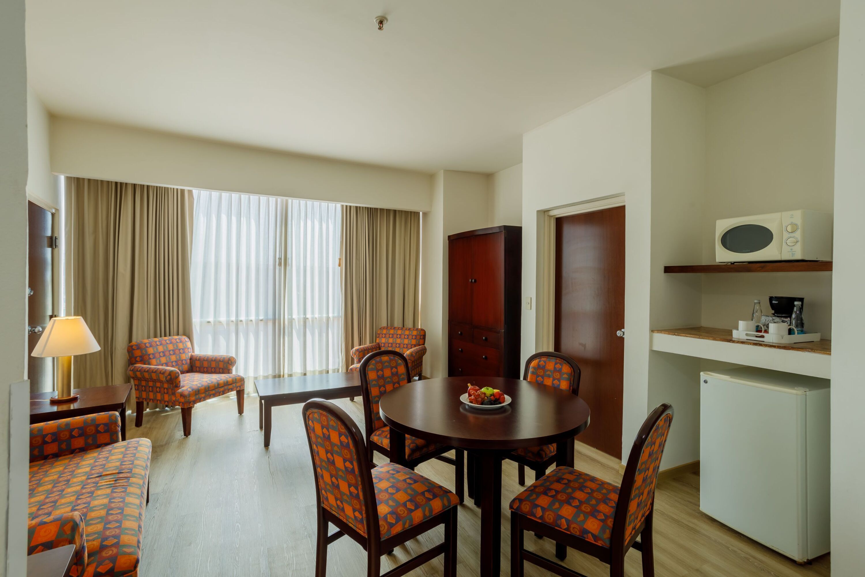 A hotel room with a dining table, four chairs, a sofa, a lamp, a microwave, and a refrigerator.