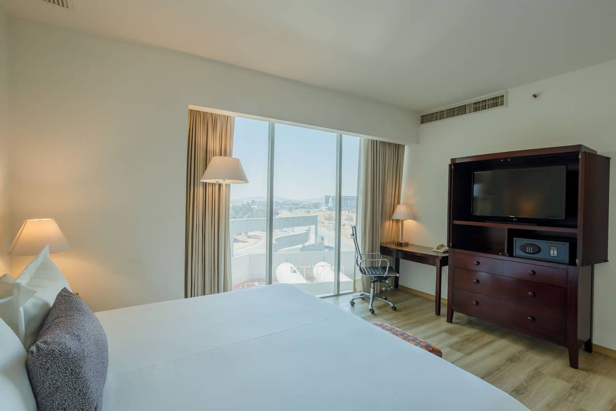 A hotel room with a large bed, a desk with a chair, a television, and a view of the city through large windows.