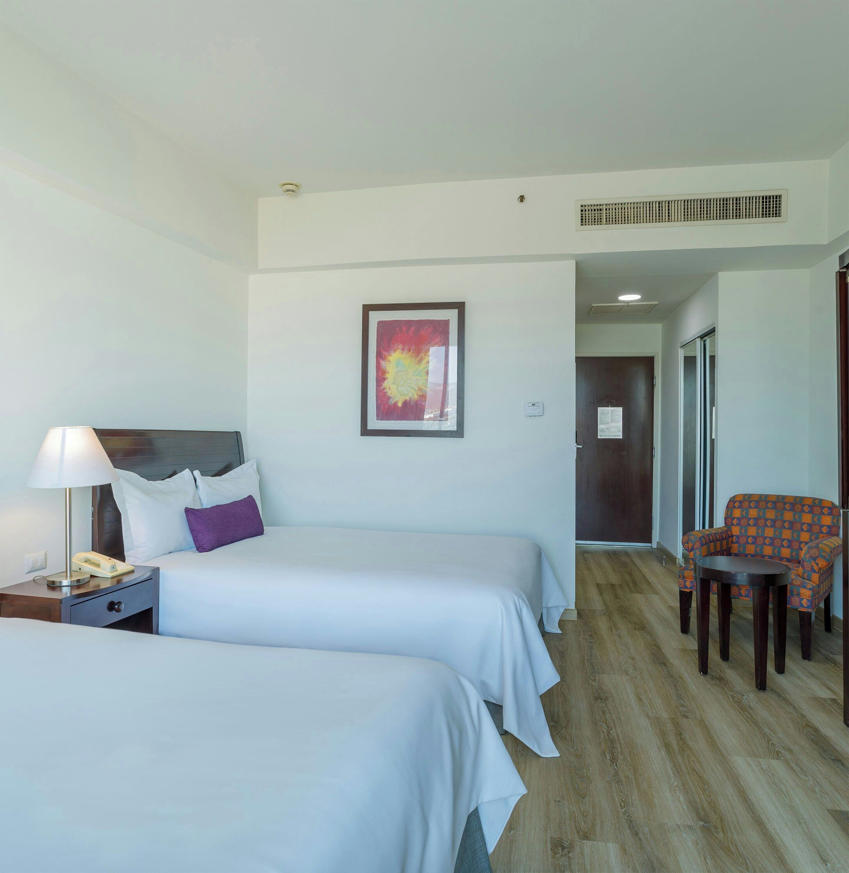A hotel room with two beds, a desk with a lamp, a chair, and a framed picture on the wall.