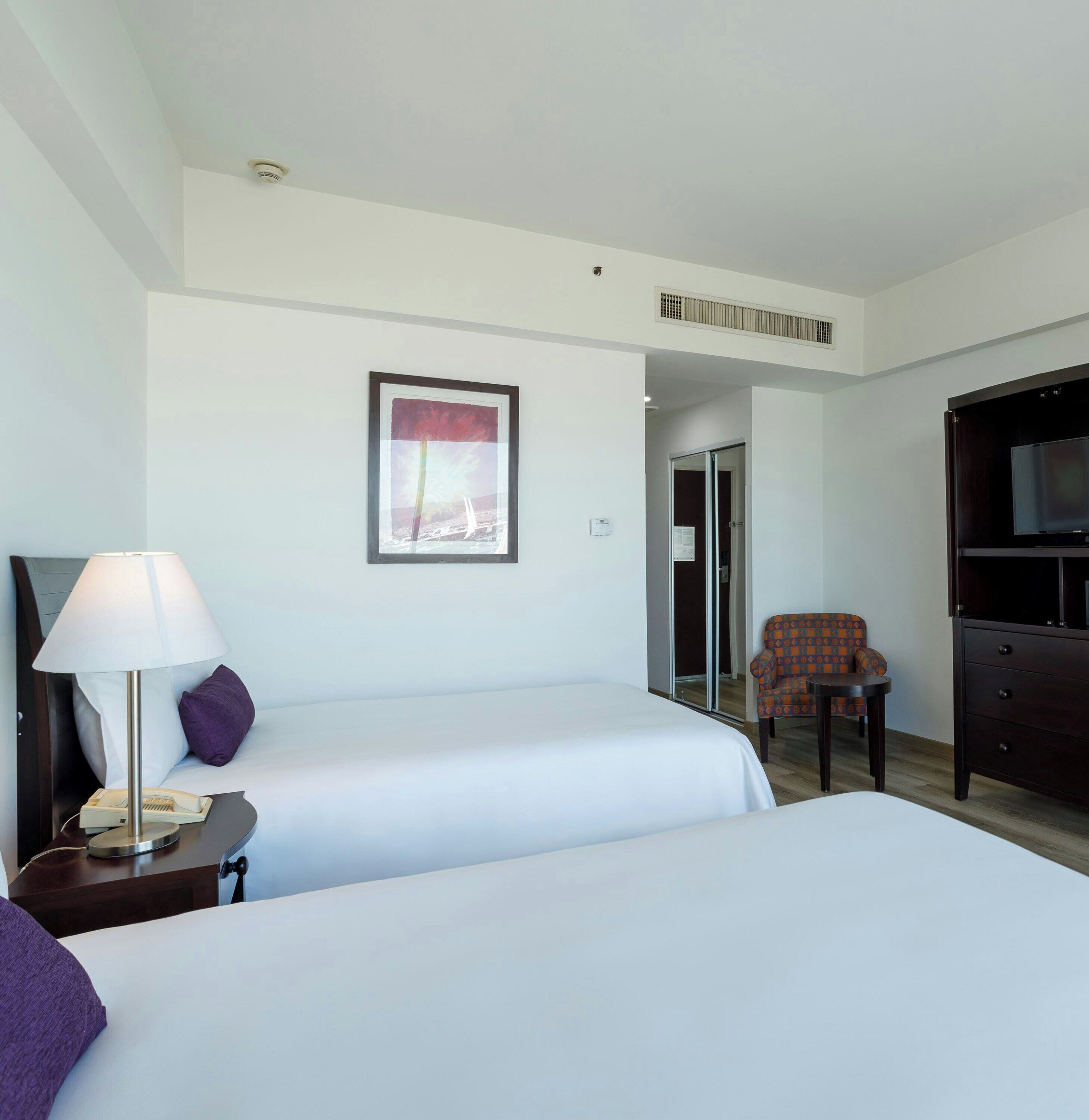 A hotel room with two beds, a desk with a lamp, a chair, a television, and a framed picture on the wall.