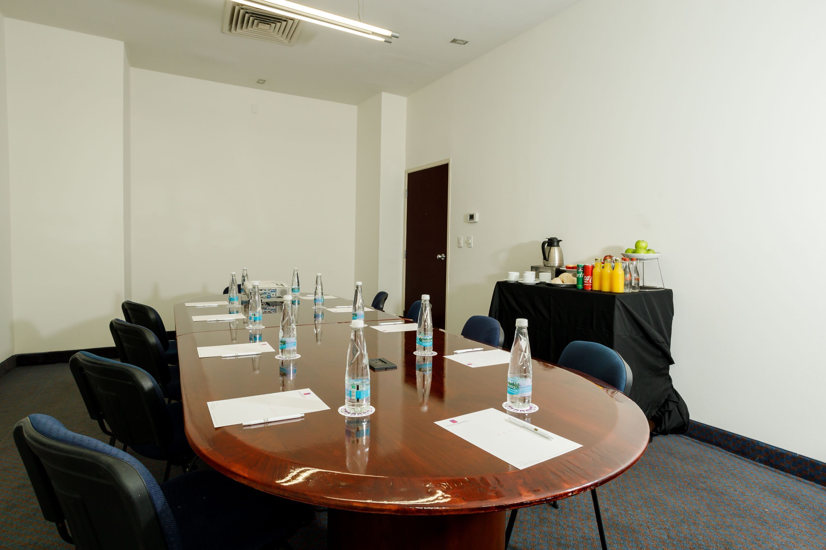 A conference room with a large oval table, black chairs, water bottles, papers, a coffee station, and a door in the background.