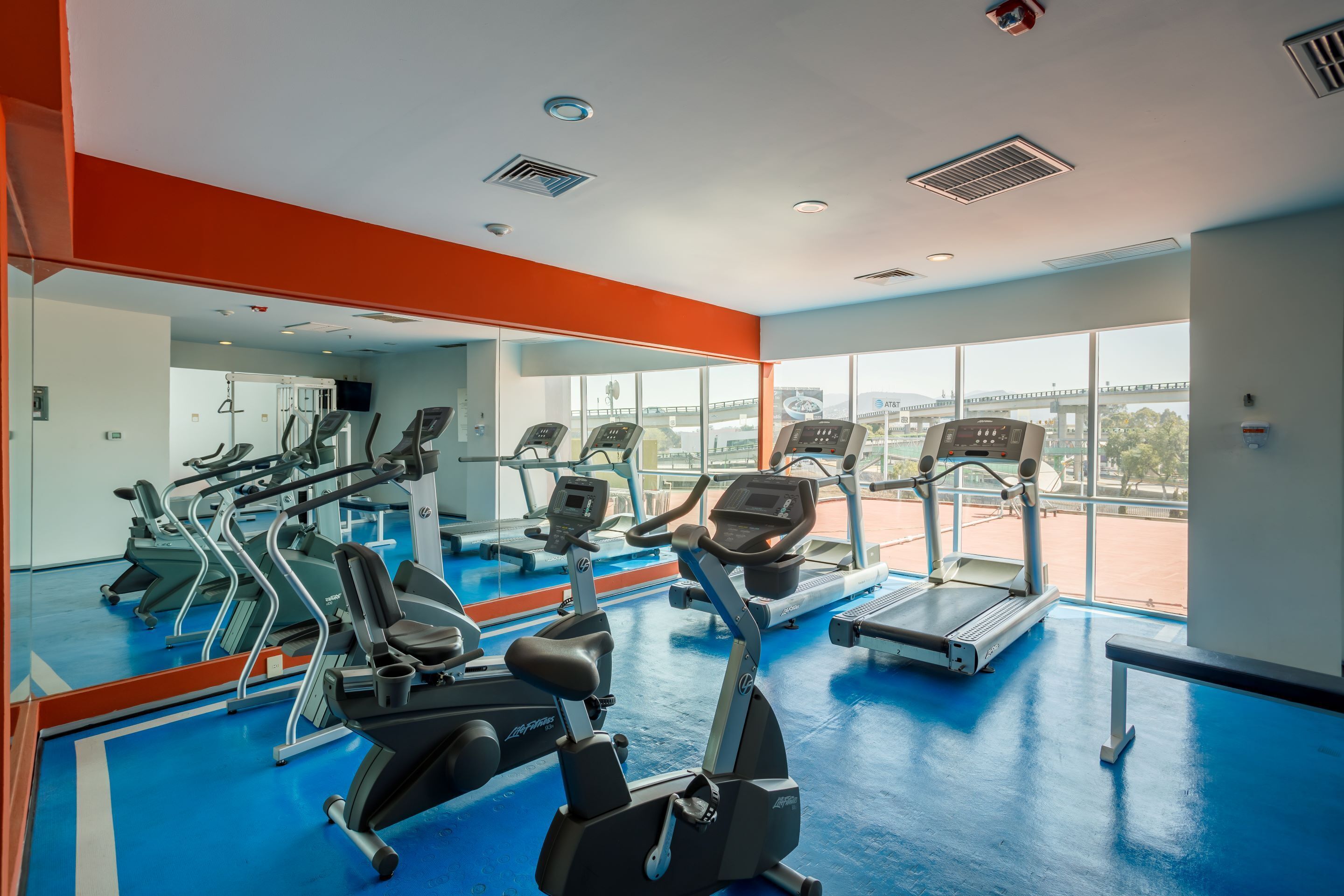 Ein Fitnessstudio mit verschiedenen Trainingsgeräten, großen Fenstern und einem blauen Boden.