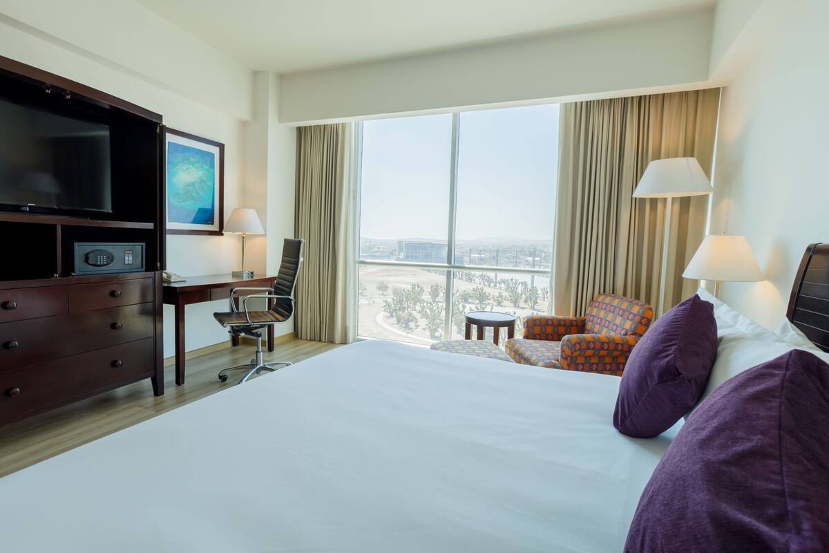 A hotel room with a large bed, a desk with a chair, a television, and a view of a cityscape through large windows.