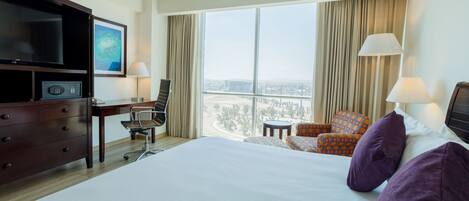 A hotel room with a large bed, a desk with a chair, a television, and a view of a cityscape through large windows.