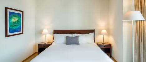 A neatly made bed with white linens, two bedside tables with lamps, a single pillow, and a framed artwork on the wall.