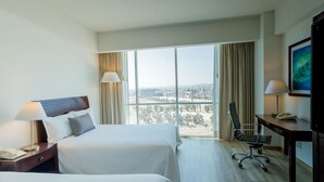 Habitación de hotel con dos camas, un escritorio, una silla y un ventanal con vistas a la ciudad.