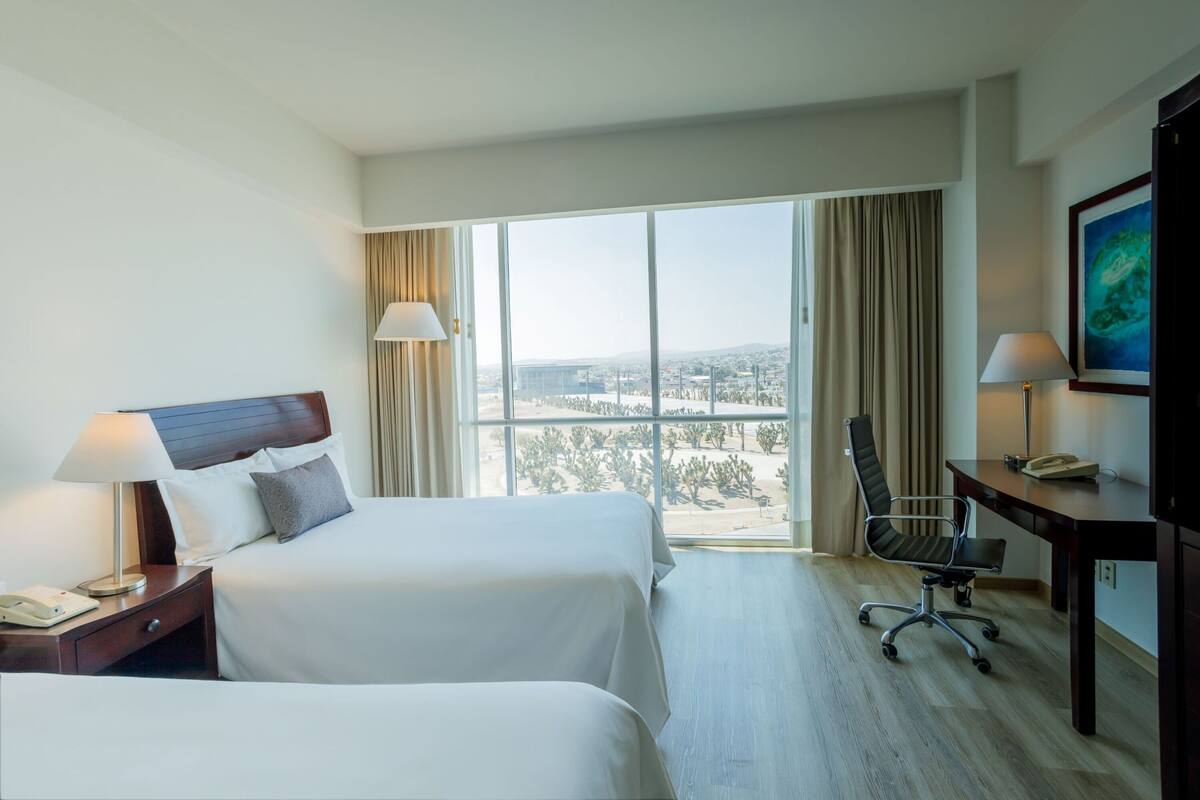 A hotel room with two beds, a desk, a chair, and a large window with a view of the city.