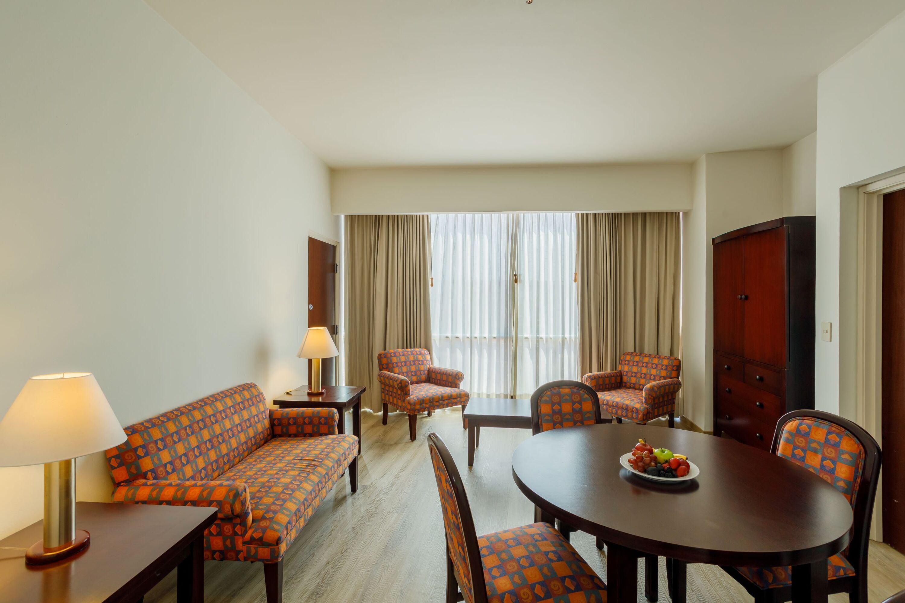 A hotel room with a sofa, two armchairs, a dining table with chairs, a lamp, and a window with curtains.