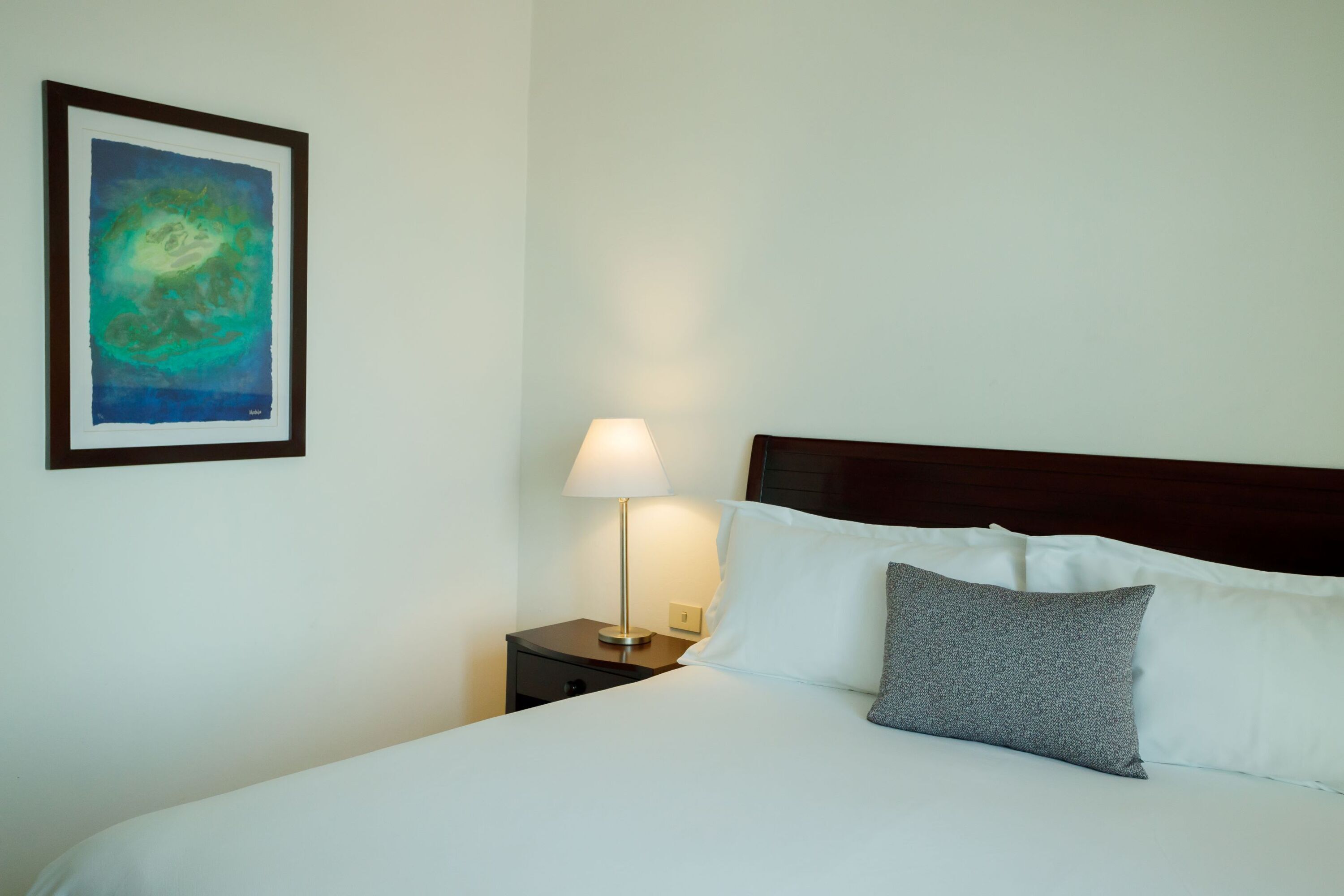 Una cama bien hecha con almohadas blancas y grises, un cuadro abstracto enmarcado en la pared y una lámpara de noche sobre una mesita de madera.