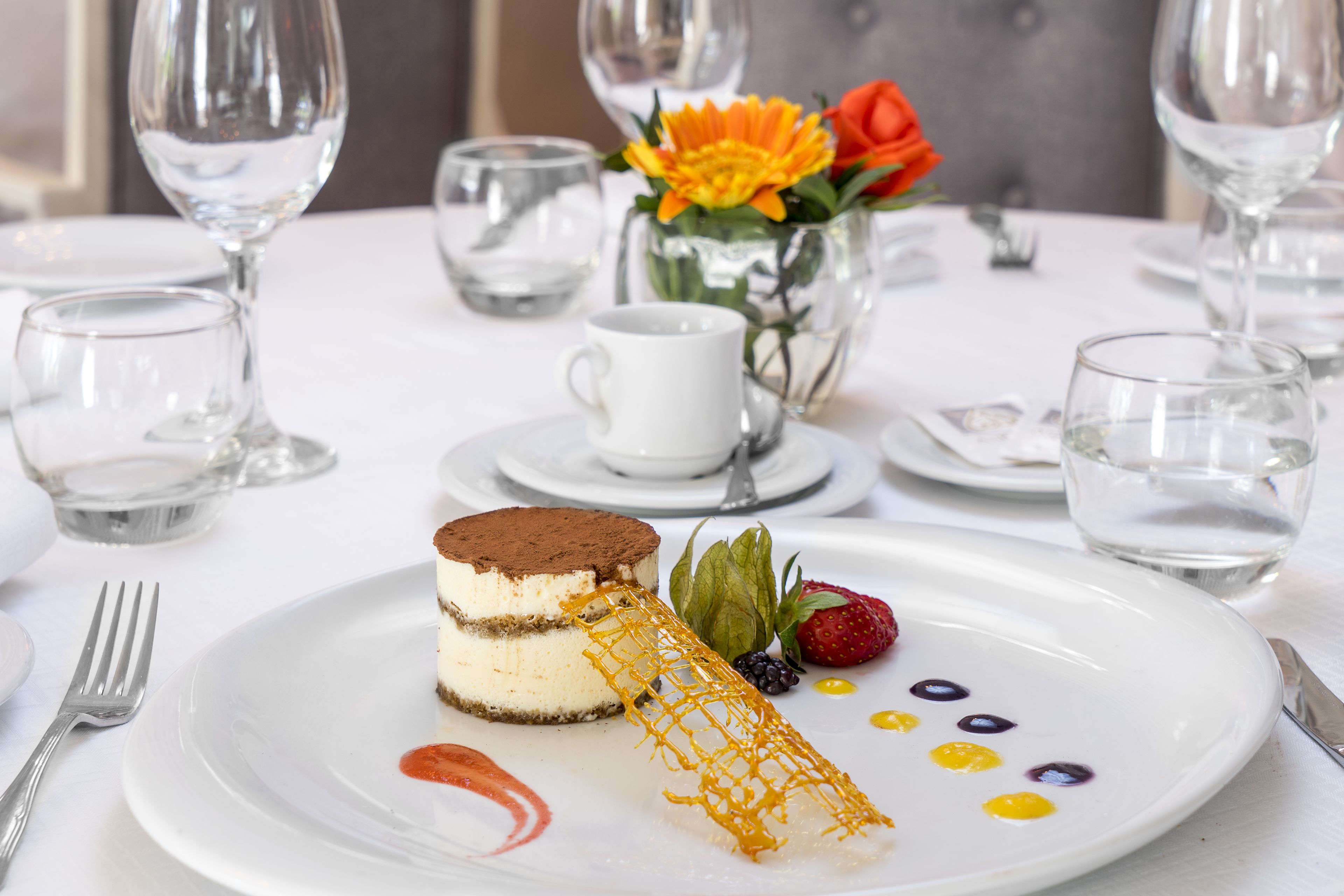 A dessert plate with a layered cake and decorative elements, accompanied by a cup and a vase with flowers.