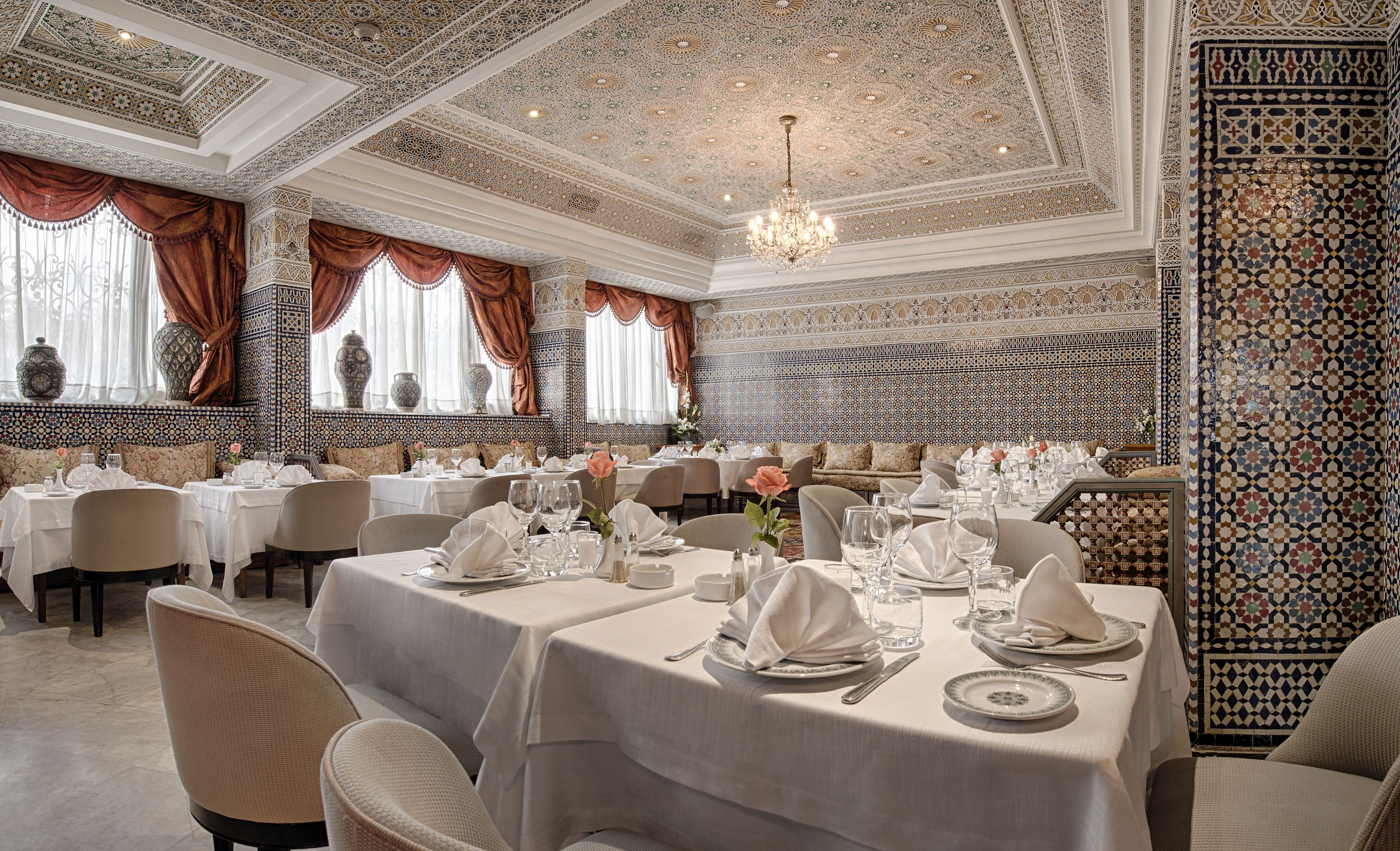 A luxurious dining room with ornate ceiling patterns, elegant table settings, and traditional Moroccan decor.