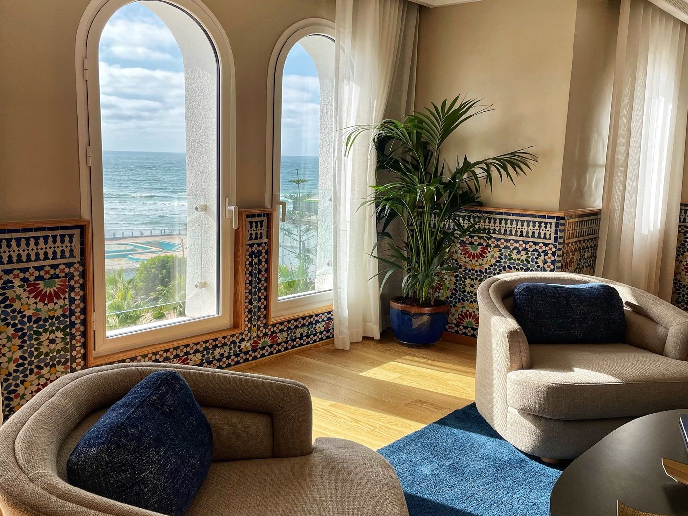 Quarto com vista para o oceano, duas poltronas, um vaso de planta e parede revestida com padrão geométrico.