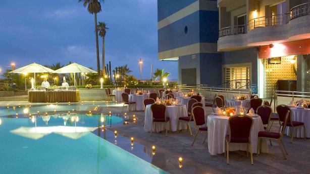 An outdoor dining area with tables set for a meal, a pool, and a building with balconies. - Club Val D Anfa (Casablanca)