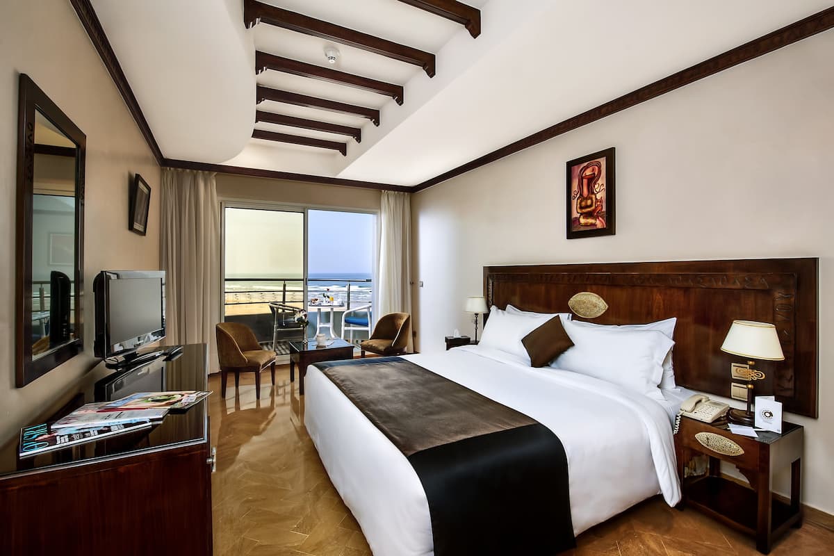 A hotel room with a large bed, a TV, a desk with a chair, a balcony view, and a wooden ceiling.