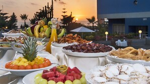 A buffet table with various fruits and pastries, set against a sunset view with palm trees and a building in the background. - Club Val D Anfa (Casablanca)